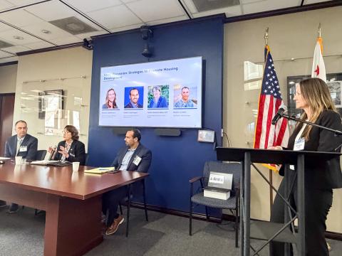 Panel discussion with four speakers and a woman at a podium, U.S. flags in background.